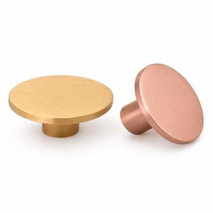 May include: Two circular cabinet knobs with a brushed metal finish. One knob features a warm gold tone, while the other displays a soft copper hue. Both items have a flat, round face and a cylindrical base, showcasing a simple, functional design.