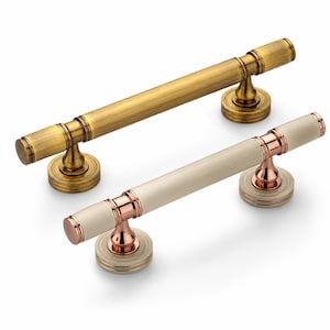 May include: Two cylindrical cabinet pulls with circular mounting bases. One pull features a brushed antique brass finish throughout. The second pull combines a smooth cream-coloured centre bar with polished rose gold accents on the ends and base.