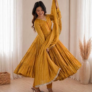 Könnte beinhalten: Senfgelber Anarkali-Anzug mit fließender Silhouette. Das langärmlige Kleid hat vertikale weiße Streifen und einen passenden Dupatta. Das Outfit umfasst eine eng anliegende Hose und verzierte Absätze. Der Gesamtstil ist elegant und traditionell.