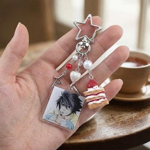 Op de afbeelding: Een zilveren sleutelhanger met een stervormige bovenkant, met een vierkante bedel met een anime-karakterillustratie, een kleine cakebedel en decoratieve kralen. De sleutelhanger bevat een hartbedel en een sterbedel.