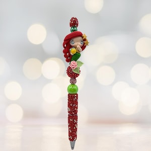 Puede incluir: Bol&iacute;grafo decorativo con dise&ntilde;o de sirena. El bol&iacute;grafo tiene un cuerpo rojo con incrustaciones de joyas y est&aacute; rematado con una figura de sirena esculpida con pelo rojo, un pez amarillo y una estrella. Incluye cuentas verdes y rosas.