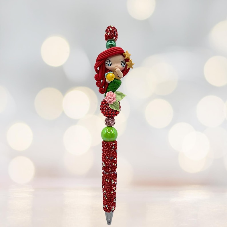 Puede incluir: Bol&iacute;grafo decorativo con dise&ntilde;o de sirena. El bol&iacute;grafo tiene un cuerpo rojo con incrustaciones de joyas y est&aacute; rematado con una figura de sirena esculpida con pelo rojo, un pez amarillo y una estrella. Incluye cuentas verdes y rosas.