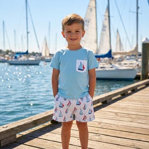 Puede incluir: Una camiseta de manga corta de color azul claro con un bolsillo blanco en el pecho que presenta un pequeño gráfico de velero, combinada con pantalones cortos blancos decorados con un patrón de pequeños veleros rojos y azules sobre un muelle.
