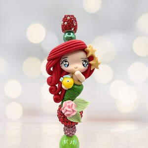 Puede incluir: Una caprichosa figura de sirena con cabello rojo, cola verde y un pez amarillo. La figura est&aacute; adornada con accesorios en forma de estrella y se asienta sobre una base decorativa con detalles rojos y rosas. La base mide aproximadamente 10 cm de altura.