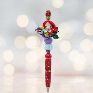 Puede incluir: Un bol&iacute;grafo decorativo con un dise&ntilde;o de sirena. El bol&iacute;grafo tiene un cuerpo rojo brillante y est&aacute; adornado con cuentas de colores y una figura de sirena. La sirena tiene el pelo rojo y monta un caballito de mar.