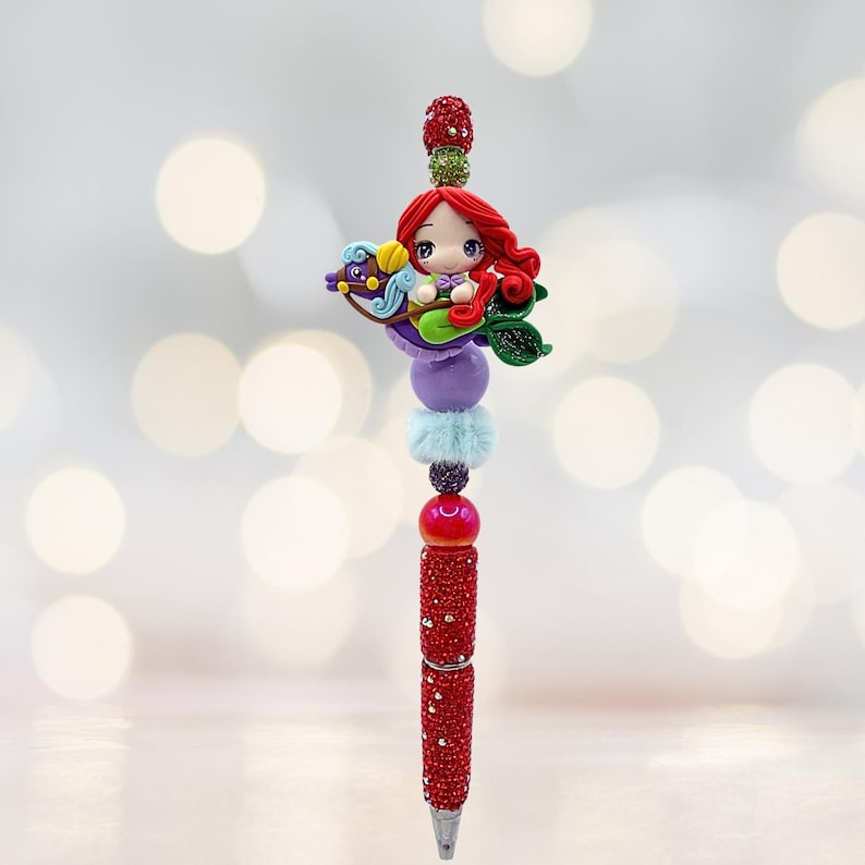 Puede incluir: Un bol&iacute;grafo decorativo con un dise&ntilde;o de sirena. El bol&iacute;grafo tiene un cuerpo rojo brillante y est&aacute; adornado con cuentas de colores y una figura de sirena. La sirena tiene el pelo rojo y monta un caballito de mar.