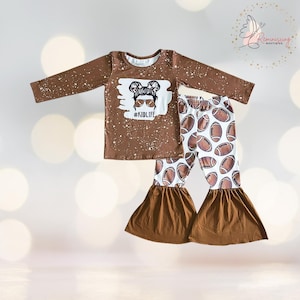 Könnte beinhalten: Ein braunes Langarmshirt mit weißem Farbspritzereffekt und einer Grafik einer Person mit Dutt, die eine Sonnenbrille in Football-Form trägt, inklusive dem Text #KIDLIFE. Dazu eine weiße Schlaghose mit einem braunen Football-Muster bedruckt.