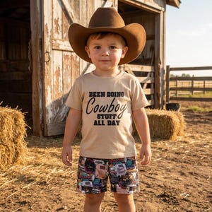 Op de afbeelding: Een beige t-shirt met de zwarte tekst BEEN DOING Cowboy STUFF ALL DAY, gecombineerd met een short met diverse kleurrijke grafische ontwerpen en een bruine vilten cowboyhoed. De outfit wordt gedragen door een jong kind dat buiten staat.