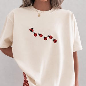 以下が含まれることがあります： アイボリー色のTシャツ。赤いテントウムシのグラフィックが斜めに並んでいます。半袖のシャツはクルーネックでゆったりとしたフィット感です。「Ivory」の文字が画像の下部に白いサンセリフ体で印刷されています。モデルは赤と白のチェック柄のショートパンツを着用しています。