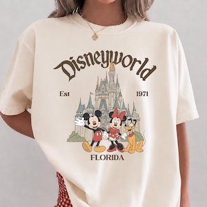 May include: Ivory-colored t-shirt featuring a vintage-style Disney World graphic. The design includes Mickey Mouse, Minnie Mouse, and Pluto in front of a castle, with the text "Disneyworld", "Est 1971", and "Florida".