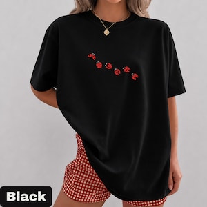 以下が含まれることがあります： クルーネックと半袖の黒いTシャツ。胸には赤いテントウムシのグラフィックが並んでいます。画像の下部にある黒い長方形には「Black」という文字が表示されています。