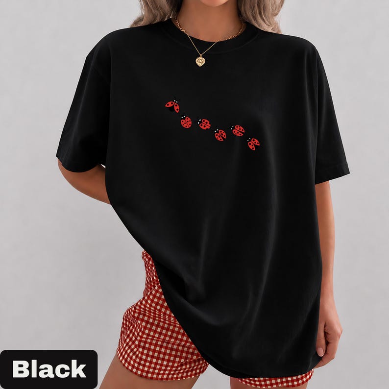 以下が含まれることがあります： クルーネックと半袖の黒いTシャツ。胸には赤いテントウムシのグラフィックが並んでいます。画像の下部にある黒い長方形には「Black」という文字が表示されています。