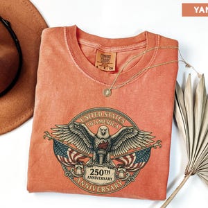 May include: A yam-colored t-shirt featuring a graphic of an eagle with outstretched wings, flags, and the text "UNITED STATES OF AMERICA 250TH ANNIVERSARY". The shirt has a round neckline and short sleeves.