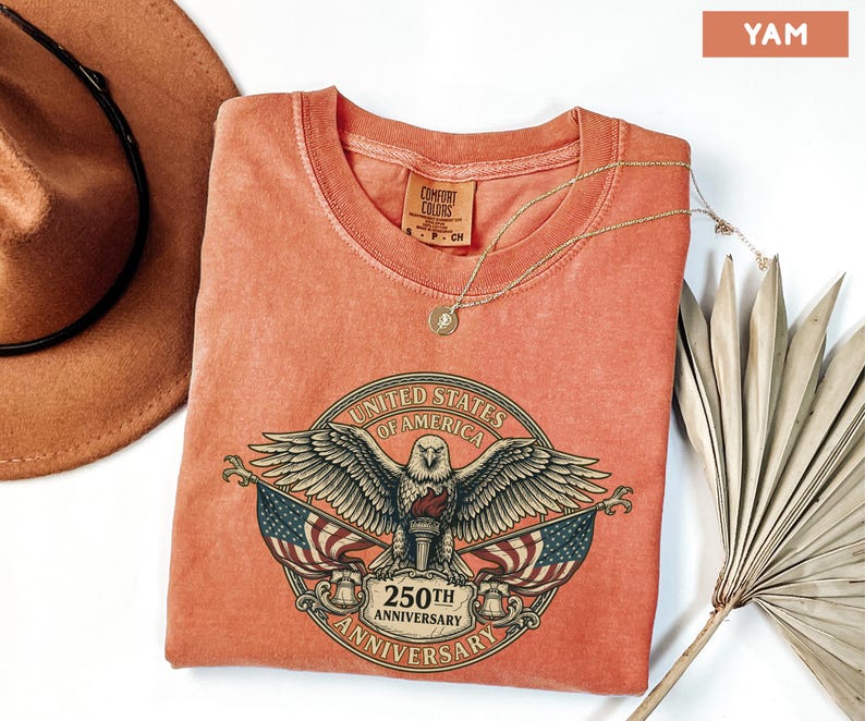 May include: A yam-colored t-shirt featuring a graphic of an eagle with outstretched wings, flags, and the text "UNITED STATES OF AMERICA 250TH ANNIVERSARY". The shirt has a round neckline and short sleeves.