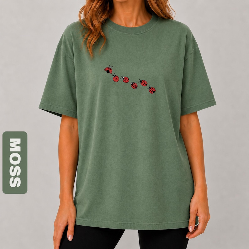 以下が含まれることがあります： モスグリーンのTシャツ。クルーネックと半袖。胸には6匹の赤いテントウムシのデザインが刺繍されています。「MOSS」の文字が左側の緑色の長方形に縦にプリントされています。