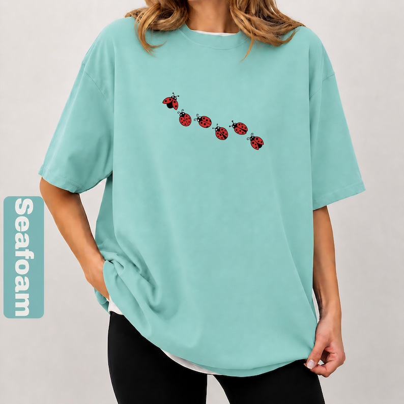 以下が含まれることがあります： 白いアンダーシャツを着た、シーフォームグリーンのTシャツ。黒い水玉模様と触角が付いた、6匹の赤いテントウムシのイラストが描かれています。ゆったりとしたフィット感のTシャツです。