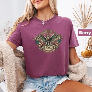 May include: Berry-colored t-shirt featuring a detailed graphic of an eagle with outstretched wings, the American flag, and the text "UNITED STATES OF AMERICA 250TH ANNIVERSARY". The shirt has short sleeves and a crew neck.