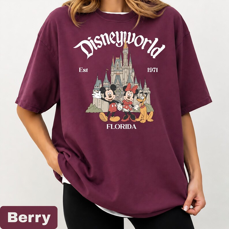 May include: Berry-colored t-shirt featuring the text "Disneyworld" above a graphic of Cinderella's castle and Mickey, Minnie, and Pluto. The shirt also includes the text "Est 1971" and "Florida".
