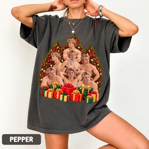 May include: A charcoal gray short-sleeve t-shirt featuring a graphic print of multiple images of a shirtless man arranged in a Christmas tree shape, decorated with red and gold ornaments, and surrounded by colorful wrapped gift boxes.