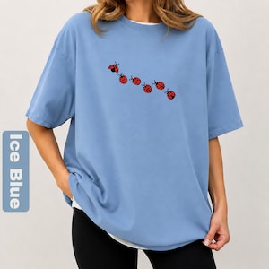 以下が含まれることがあります： 白のクルーネックと半袖の、アイスブルーのTシャツ。6匹の赤いテントウムシのデザインが並んでいます。ゆったりとしたフィット感で、柔らかい素材で作られています。