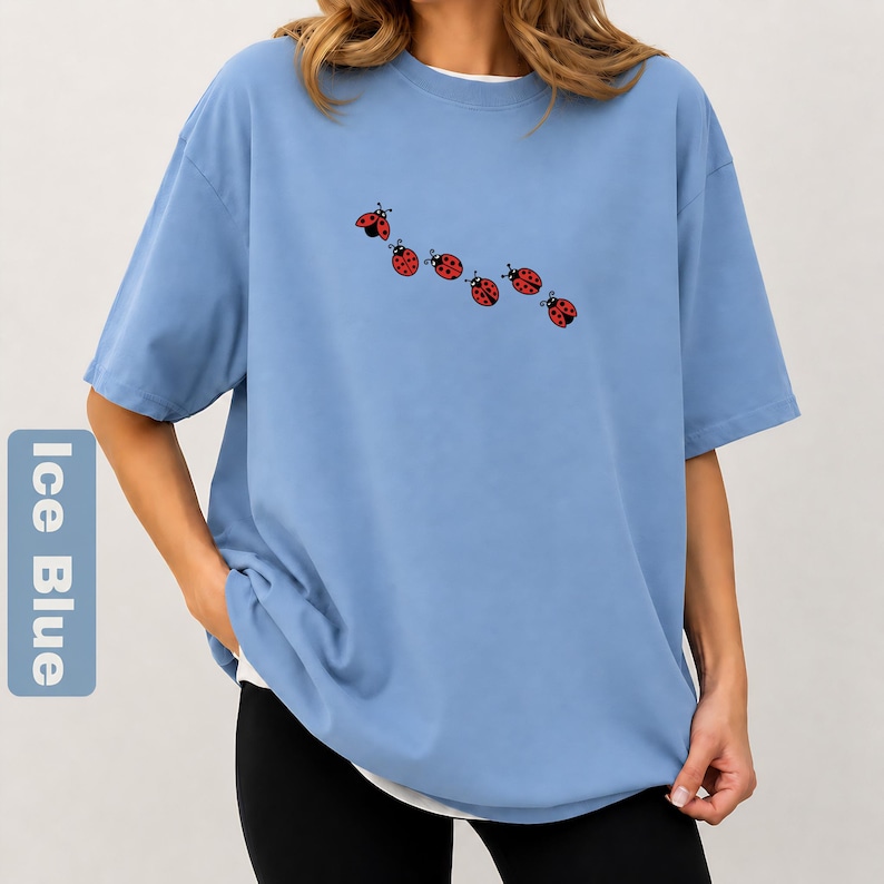 以下が含まれることがあります： 白のクルーネックと半袖の、アイスブルーのTシャツ。6匹の赤いテントウムシのデザインが並んでいます。ゆったりとしたフィット感で、柔らかい素材で作られています。