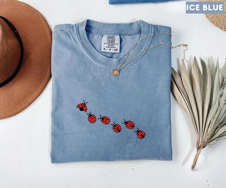 May include: Ice blue Comfort Colors t-shirt with a crew neck and a ladybug design. The ladybugs are red with black spots and are arranged in a diagonal line. A gold necklace with a pendant is draped across the shirt.