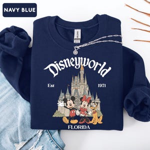 May include: Navy blue sweatshirt with "Disneyworld" text and a graphic of Mickey Mouse, Minnie Mouse, and Pluto in front of a castle. The text "Est 1971" and "Florida" are also printed on the sweatshirt. A silver necklace is draped across the top.
