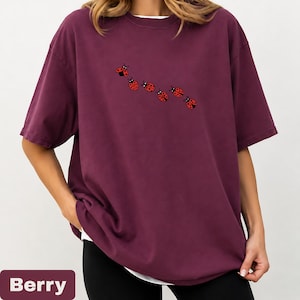 以下が含まれることがあります： ベリー色のオーバーサイズのTシャツと白いインナー。7匹の赤いテントウムシのデザインが並んでいます。「Berry」の文字が画像の下部の長方形のボックスに表示されています。