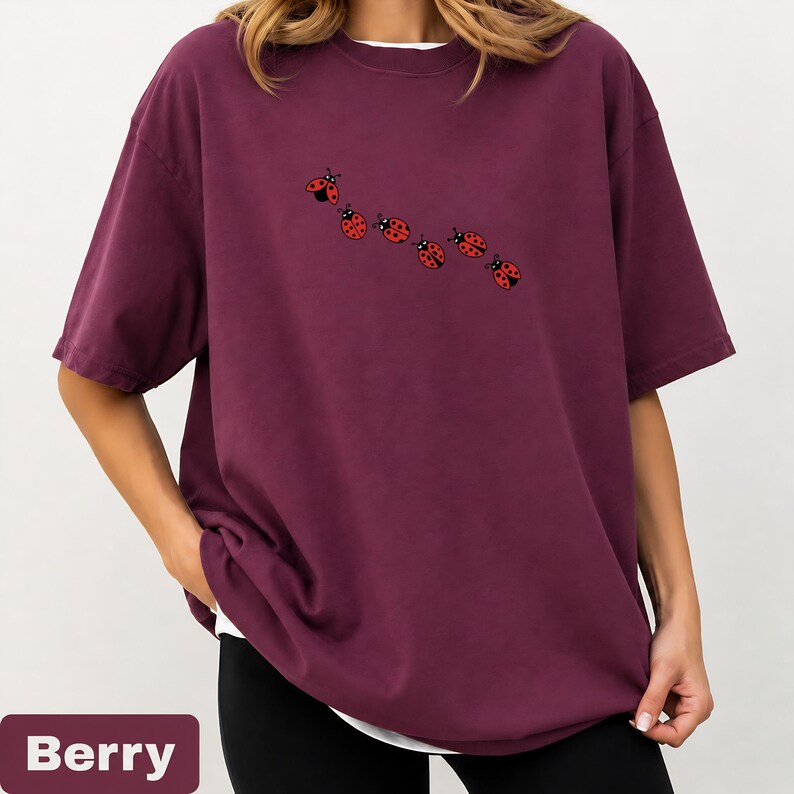 以下が含まれることがあります： ベリー色のオーバーサイズのTシャツと白いインナー。7匹の赤いテントウムシのデザインが並んでいます。「Berry」の文字が画像の下部の長方形のボックスに表示されています。