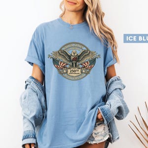 May include: Ice blue t-shirt featuring a detailed emblem with an eagle, American flags, and the text "250th Anniversary". The emblem is in a circular design with the words "United States of America".