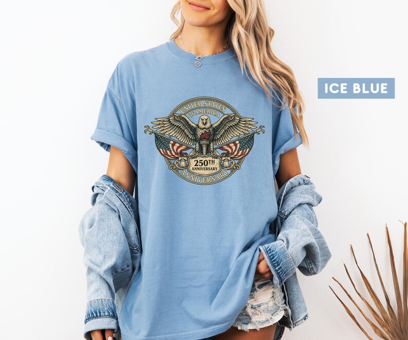 May include: Ice blue t-shirt featuring a detailed emblem with an eagle, American flags, and the text "250th Anniversary". The emblem is in a circular design with the words "United States of America".