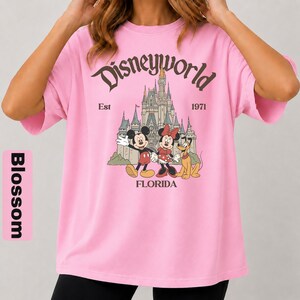 May include: A light pink t-shirt featuring a graphic of Mickey Mouse, Minnie Mouse, and Pluto in front of Cinderella's castle. The text "Disneyworld" is arched above the castle, with "Est 1971" and "Florida" below.