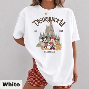 May include: White t-shirt featuring a vintage-style Disney World graphic. The design includes Mickey Mouse, Minnie Mouse, Pluto, and Cinderella's castle, with the text "Disneyworld Est 1971 Florida."