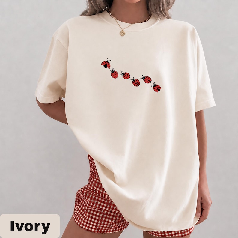 以下が含まれることがあります： アイボリー色のTシャツ。赤いテントウムシのグラフィックが斜めに並んでいます。半袖のシャツはクルーネックでゆったりとしたフィット感です。「Ivory」の文字が画像の下部に白いサンセリフ体で印刷されています。モデルは赤と白のチェック柄のショートパンツを着用しています。