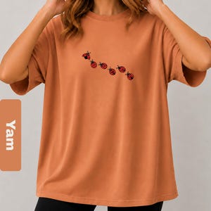 以下が含まれることがあります： 7匹の赤と黒のてんとう虫のイラストが描かれた、オレンジ色のTシャツ。半袖のシャツはゆったりとしたフィット感で、クルーネックです。「Yam」の文字が縦長の長方形のラベルにプリントされています。
