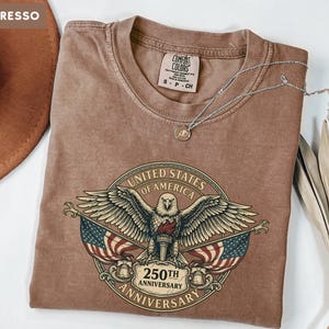 May include: Espresso-colored t-shirt featuring a graphic of an eagle, American flags, and the text "United States of America 250th Anniversary." A silver necklace with a pendant rests on the shirt. A brown hat is visible in the upper left corner.