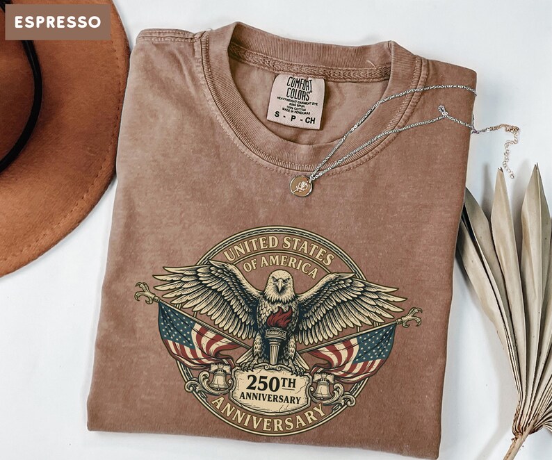 May include: Espresso-colored t-shirt featuring a graphic of an eagle, American flags, and the text "United States of America 250th Anniversary." A silver necklace with a pendant rests on the shirt. A brown hat is visible in the upper left corner.