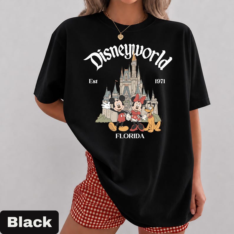 May include: Black t-shirt featuring the text "Disneyworld" above an illustration of Cinderella's castle, Mickey Mouse, Minnie Mouse, and Pluto. The text "Est 1971 Florida" is also included. The shirt is a classic crew neck.