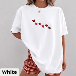 以下が含まれることがあります： 赤いテントウムシのデザインが並んだ白いTシャツ。Tシャツは半袖でゆったりとしたフィット感です。赤と白のチェック柄のショートパンツも写っています。画像の下部には「White」の文字があります。