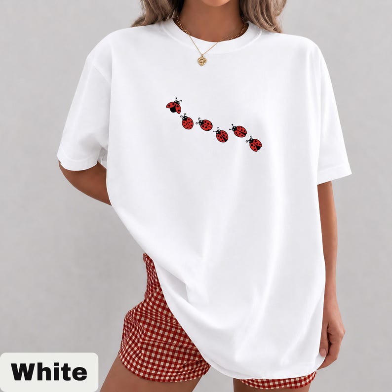 以下が含まれることがあります： 赤いテントウムシのデザインが並んだ白いTシャツ。Tシャツは半袖でゆったりとしたフィット感です。赤と白のチェック柄のショートパンツも写っています。画像の下部には「White」の文字があります。