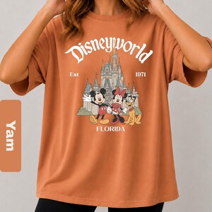 May include: Orange t-shirt with a Disney World graphic. The design features Mickey Mouse, Minnie Mouse, and Pluto in front of a castle, with the words "Disneyworld", "Florida", and "Est 1971".