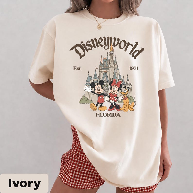 May include: Ivory-colored t-shirt featuring a vintage-style Disney World graphic. The design includes Mickey Mouse, Minnie Mouse, and Pluto in front of a castle, with the text "Disneyworld", "Est 1971", and "Florida".