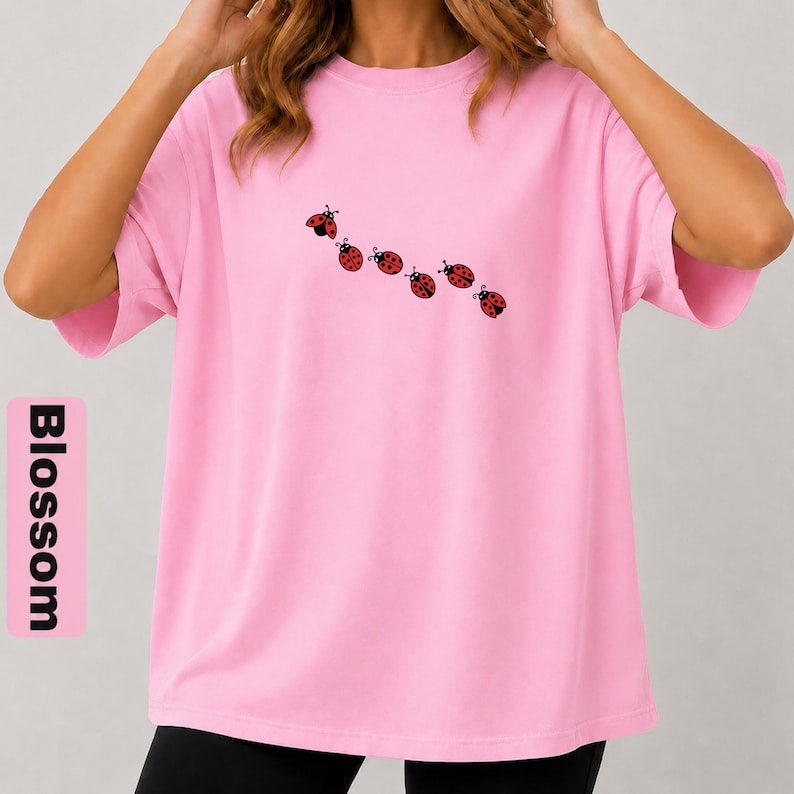 以下が含まれることがあります： 黒と赤のてんとう虫のイラストが7つ並んだ、明るいピンク色のTシャツ。「Blossom」の文字がシャツの左側に縦にプリントされています。