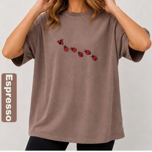 以下が含まれることがあります： 赤色のてんとう虫のグラフィックが並んだ、エスプレッソ色のTシャツ。半袖のシャツはゆったりとしたフィット感です。「Espresso」の文字が、画像の左側にある茶色の長方形に縦に印刷されています。
