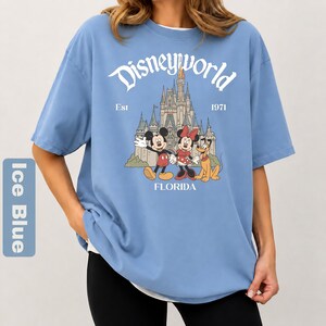 May include: Ice blue t-shirt with a Disneyworld graphic. The graphic features Mickey Mouse, Minnie Mouse, and Pluto in front of a castle. The text "Disneyworld Est 1971 FLORIDA" is printed above and below the characters.