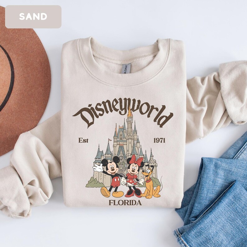 May include: A sand-colored sweatshirt featuring a Disney World graphic. The design includes Mickey Mouse, Minnie Mouse, and Pluto in front of a castle, with the words "Disneyworld", "Est 1971", and "Florida". The sweatshirt is folded on a white surface.