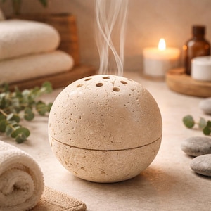 Peut inclure: Diffuseur d'huiles essentielles sphérique beige avec une surface texturée et de petits trous sur le dessus, dégageant de la vapeur. Le diffuseur est divisé en deux moitiés. L'arrière-plan comprend une bougie allumée, des serviettes et des petites pierres.