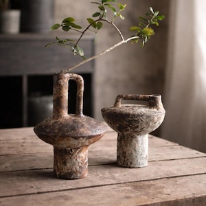 Peut inclure: Deux vases bruns patinés avec des poignées originales sur une table en bois. L'un a une poignée haute et ouverte, l'autre une poignée plus courte et fermée. Une branche de verdure ajoute une touche naturelle.