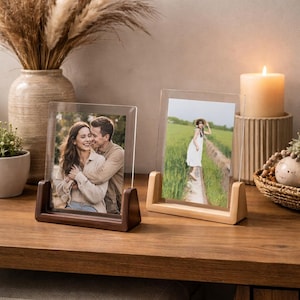 Peut inclure: Deux cadres photo en acrylique transparent avec des socles en bois. L'un a une base marron foncé et affiche une photo d'un couple. L'autre a une base en bois clair et montre une personne dans un champ. Une bougie allumée et des objets décoratifs sont en arrière-plan.