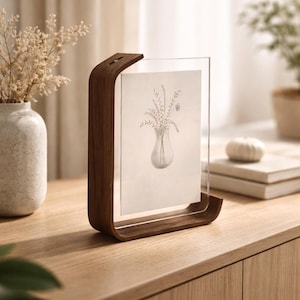 Puede incluir: Un marco de fotos de madera con una parte frontal de acrílico transparente muestra una ilustración botánica de flores en un jarrón. El marco tiene un diseño curvo y se apoya en una superficie de madera. Un jarrón y libros están en el fondo.